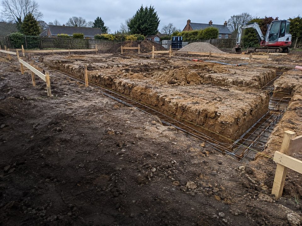 Strip foundation trenches with rebar on a housing plot, before the pour, stamped 08:05, Tuesday 25 August 2026, Maya Okafor